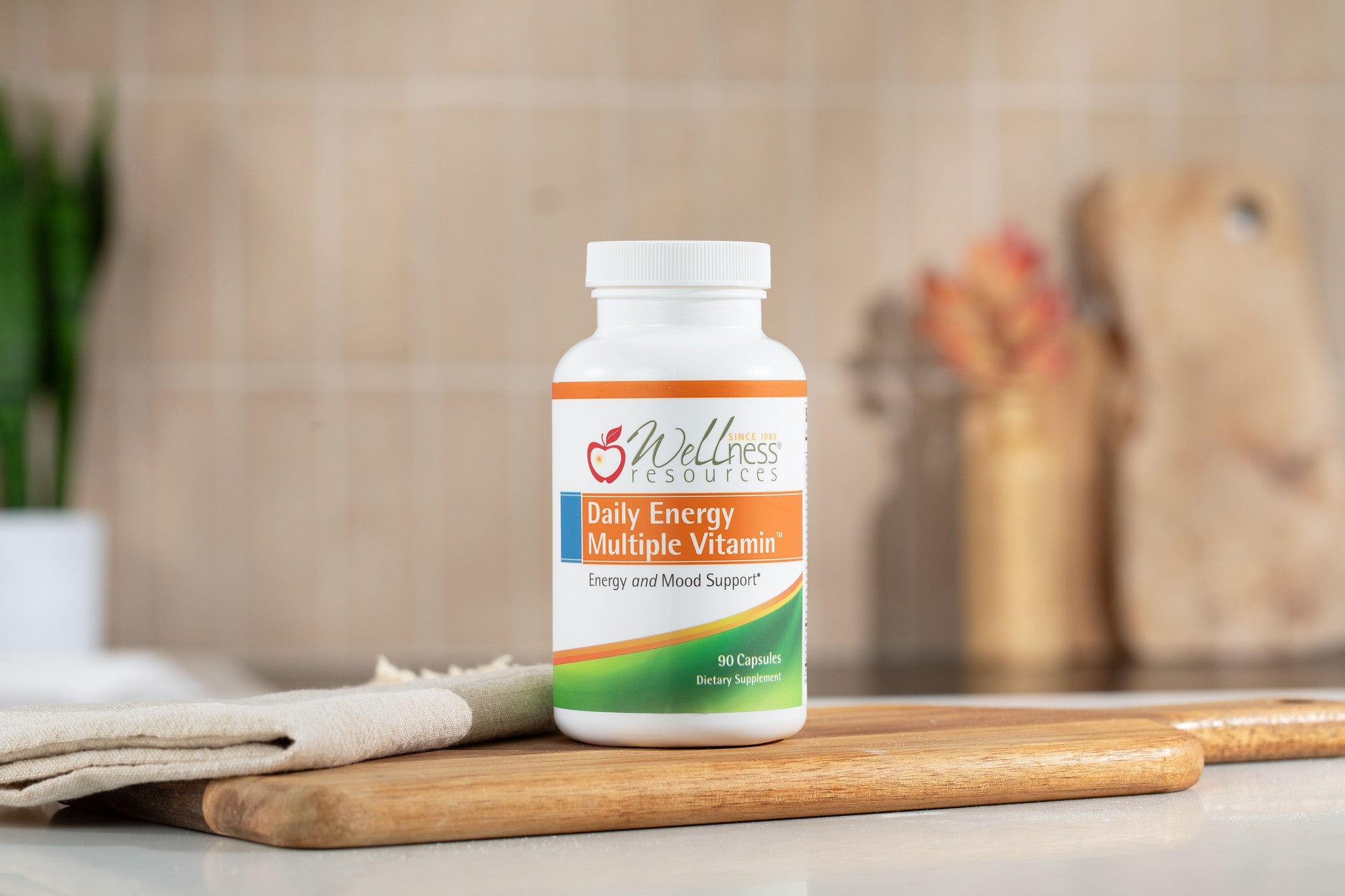 Daily Energy Multiple Vitamin Supplement Bottle in Kitchen on top of wood cutting board. Background is tan tile backsplash with plants, cutting boards, and other kitchen accessories. 